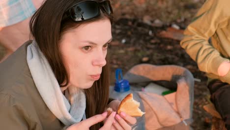 Young-brunette-woman-eating-bread-and-meat-fried-on-fire.-Camping-trip.