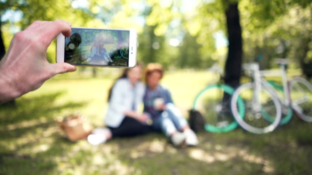 Freund-ist-zwei-hübsche-Mädchen-sitzen-auf-Rasen-im-Park-mit-Handy-in-der-hand-halten-Fokus-auf-Smartphone-Bildschirm-dann-auf-Menschen-fotografieren.-Porträt-und-Technologie-Konzept.
