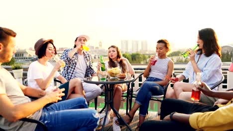 Fröhliche-Junge-Menschen-feiern-Urlaub-am-Dach-zu-trinken-und-klirrende-Flaschen-und-Gläser,-dann-reden-und-lachen.-Schöne-Aussicht-auf-moderne-Stadt-ist-im-Hintergrund.