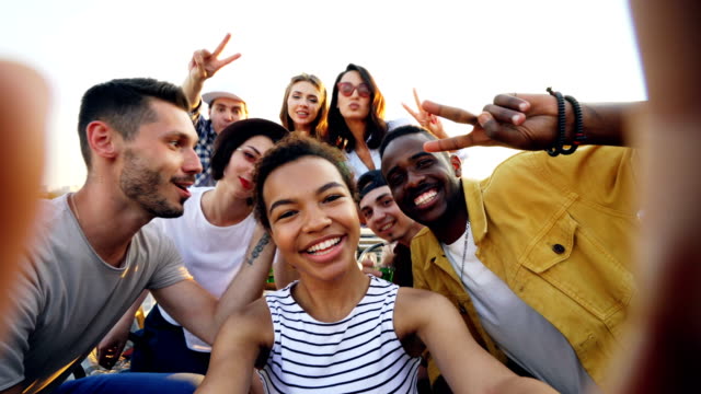 Point-of-view-shot-of-African-American-girl-holding-camera-and-taking-selfie-with-happy-friends-at-party-on-roof.-Men-and-women-are-looking-at-camera,-posing-and-laughing.