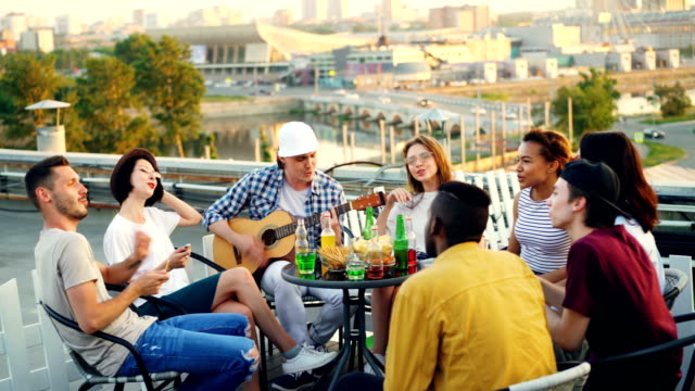 Young-people-are-having-fun-on-rooftop-playing-the-guitar,-singing,-chatting-and-laughing-sitting-at-table-outdoors.-Joy,-music,-youth-and-friendship-concept.