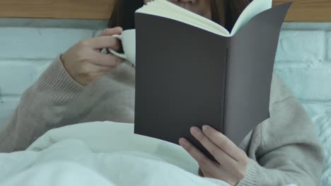 Beautiful-asian-woman-enjoying-drinking-warm-coffee-and-reading-book-on-bed-in-her-bedroom.Asia-female-wearing-comfortable-sweater-holding-a-book-and-cup-of-coffee.lifestyle-asia-woman-at-home-concept