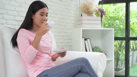 Cheerful-asian-young-woman-drinking-warm-coffee-or-tea-enjoying-it-while-sitting-in-her-living-room-at-home.-Attractive-happy-asian-woman-holding-a-cup-of-coffee.