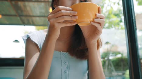 Fröhliche-Junge-Asiatin-trinken-warmen-Kaffee-oder-Tee-genießen-sie-im-Café-sitzen.-Attraktive-glücklich-asiatische-Frau-hält-eine-Tasse-Kaffee.