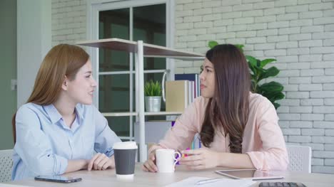 Two-young-Asian-women-college-students-or-coworkers-drinking-coffee-and-talking-in-office,-diverse-group.-Casual-business,-freelance-work-at-cafe,-social-meeting-concept