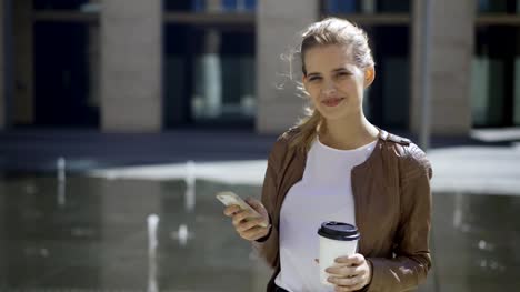 Beautiful-blonde-woman-with-disposable-coffee-cup-and-cell-phone-in-her-hands-posing-near-fountain-in-street-and-smiling-happily-at-camera,-medium-shot