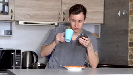 Young-man-with-a-phone-eats-at-a-table-in-the-kitchen