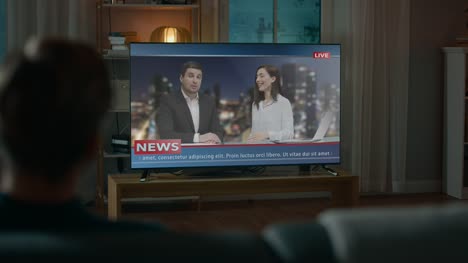 Man-Watches-Late-Night-News-Show-on-TV-while-Sitting-on-a-Couch-at-Home-in-the-Evening.-Two-Presenters-Talk-and-Joke-on-TV.-Cozy-Living-Room-with-Warm-Lights.-Over-the-Shoulder-Shot.