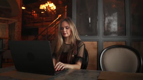 Young-woman-sitting-in-coffee-shop-at-wooden-table,-drinking-coffee-and-using-smartphone.On-table-is-laptop.-Girl-browsing-internet,-chatting,-blogging.-Female-holding-phone-and-looking-on-his-screen