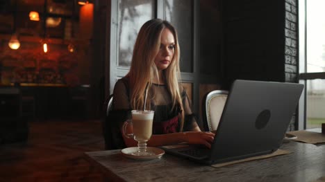 Side-view.-Young-business-woman-sitting-at-table-and-taking-notes-in-notebook.On-table-is-laptop,-smartphone-and-cup-of-coffee.On-computer-screen-graphics-and-charts.-Student-learning-online.-Blogger