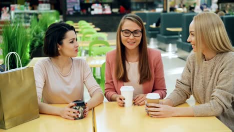 Group-of-happy-young-ladies-is-chatting-and-laughing-sitting-at-table-in-shopping-mall-cafe-with-to-go-drinks-and-socializing.-Youth-lifestyle-and-fun-concept.