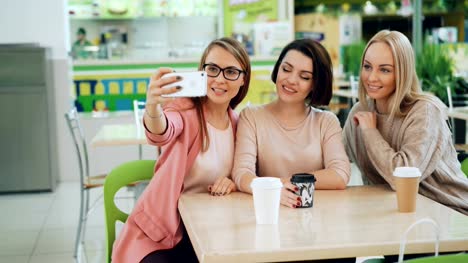 Beautiful-young-ladies-are-taking-selfie-using-smartphone-sitting-at-table-in-cafe-and-posing-with-drinks.-Friendship,-leisure-time-and-modern-technology-concept.