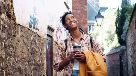 Young-black-woman-wearing-a-plaid-shirt-standing-in-an-alleyway-holding-her-coat-and-a-takeaway-coffee,-smiling-to-camera,-close-up