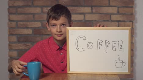 Kind-am-Schreibtisch-hält-Flipchart-mit-Schriftzug-Kaffee-auf-dem-Hintergrund-roten-Backsteinmauer