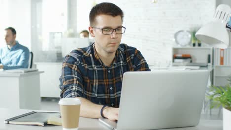 Gesicht-Front-Man-sitzt-an-seinem-kreativen-Office-Table-Working-on-His-Laptop.-Im-Hintergrund-ist-der-Mann,-der-an-ihren-Desktops-arbeitet.
