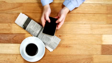 Geschäftsfrau-mit-Handy-am-Schreibtisch-mit-Zeitung-und-Kaffee-am-Tisch
