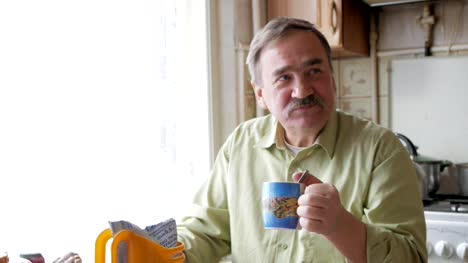 An-elderly-man-with-a-mustache-holds-a-cup-of-hot-drink-and-drinks.-He-sits-near-the-window-and-has-breakfast-at-home
