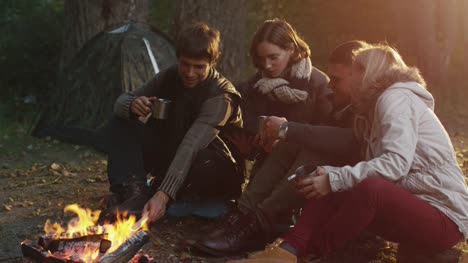 Grupo-de-personas-sentarse-en-un-bosque-junto-a-una-fogata-con-bebidas-calientes-de-termo-y-utilizar-la-tableta.
