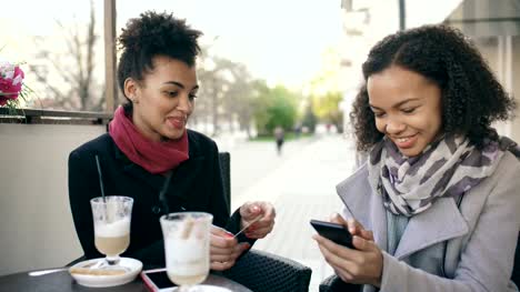 Zwei-attraktive-gemischte-Rasse-Frauen-mit-Online-Shopping-mit-Kreditkarte-und-Smartphone-beim-Sprechen-und-Kaffee-trinken-in-Straßencafé