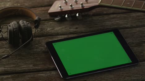 Tablet-PC-with-Green-Screen-Laying-on-Wooden-Table-next-to-Guitar-and-Headphones.-Causal-Lifestyle