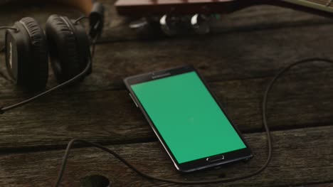 Teléfono-Android-con-pantalla-verde-en-retrato-modo-puesta-en-mesa-de-madera-junto-a-la-guitarra-y-auriculares.-Estilo-de-vida-causal