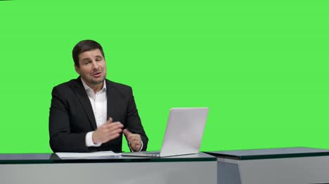 Media-broadcaster-is-sitting-at-a-table-and-talking-on-a-mock-up-green-screen-in-the-background.