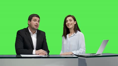 Male-and-female-broadcasters-are-sitting-at-a-table-and-talking-on-a-mock-up-green-screen-in-the-background.