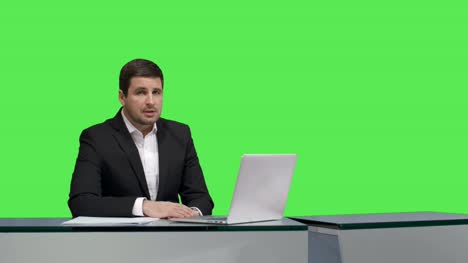 Media-broadcaster-is-sitting-at-a-table-and-talking-on-a-mock-up-green-screen-in-the-background.