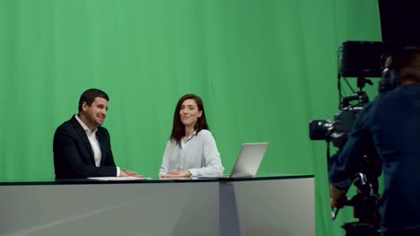 Backstage-footage-of-male-and-female-broadcasters-who-sit-at-a-table-and-talk-on-a-mock-up-green-screen-in-the-background.