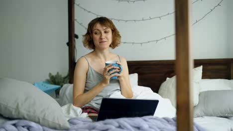 Mujer-joven-atractiva-tener-chat-de-vídeo-en-línea-con-amigos-utilizando-la-cámara-portátil-sentado-en-la-cama-en-casa