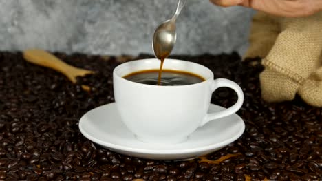 4k-Nahaufnahme-weibliche-Hand-rühren-Cup-schwarz-Kaffee---weibliche-Hand-mit-Löffel-um-zu-rühren,-frischem-Boden-schwarz-weiße-Kaffeetasse-Untertasse-neben-Kaffee-in-Bohnen,-Kaffee-Zeit