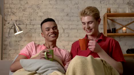 Multinational-gay-couple-sitting-on-couch-covered-with-a-warm-blanket,-holding-hands,-drink-tea,-look-at-the-camera,-laughing.-Homeliness,-romantic-evening,-background,-hugs,-happy-LGBT-family-concept.-60-fps