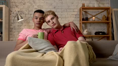 Multinationale-schwules-Paar-auf-Couch-mit-einer-warmen-Decke-bedeckt-Fernsehen,-verwenden-Sie-die-Fernbedienung,-Blick-auf-die-Kamera.-Gemütlichkeit,-romantischen-Abend,-Streicheleinheiten,-glücklich-LGBT-Familien-Konzept.-60fps