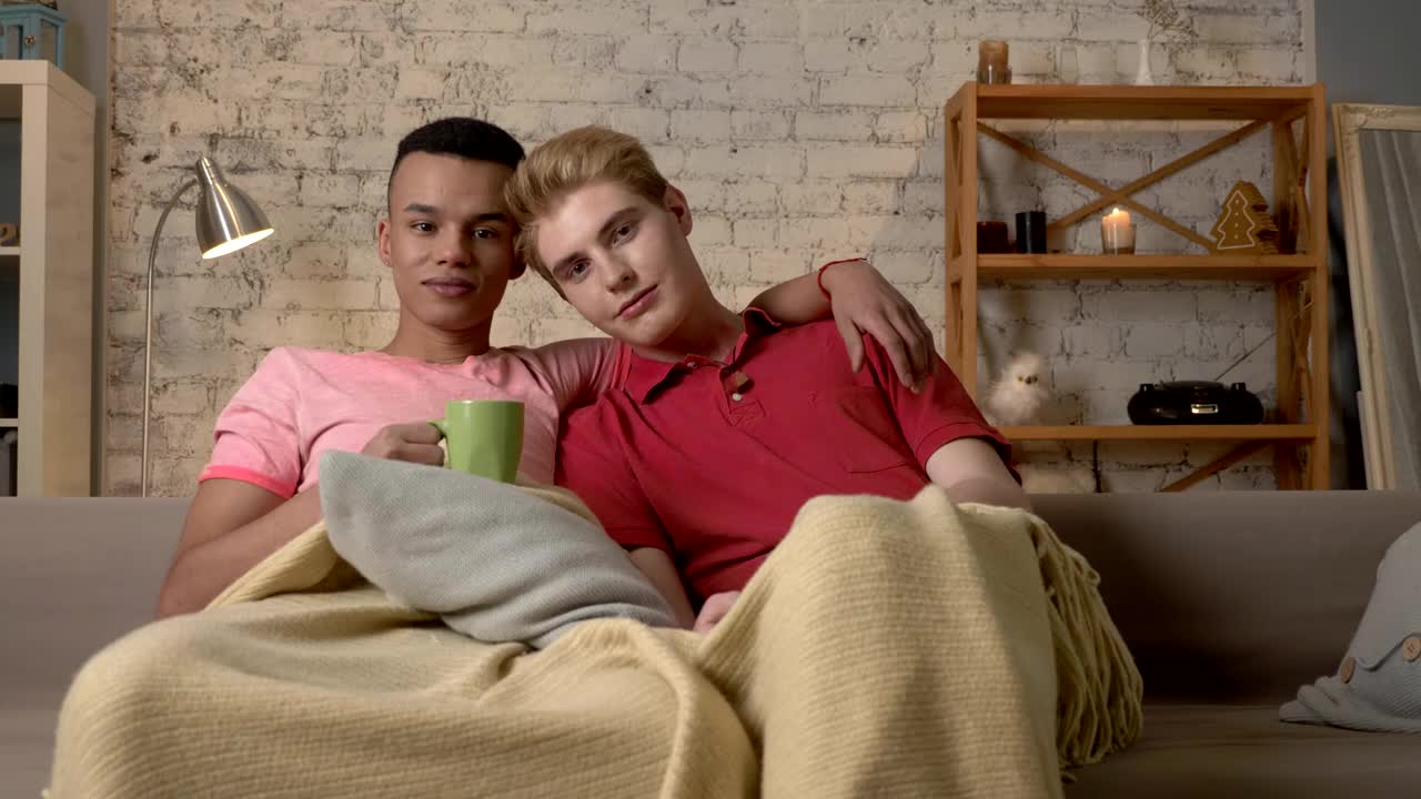 Two Gay Guys Sit On The Couch And Watch TV, Drink Hot Tea, Use The Remote  Control. LGBT Lovers, A Multinational Couple, A Happy Gay Family, A Home  Cosiness Concept. Look At