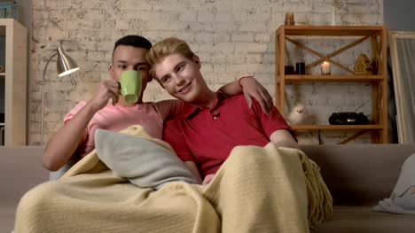 Two-gay-guys-sit-on-the-couch-and-watch-TV,-drink-hot-tea,-use-the-remote-control.-LGBT-lovers,-a-multinational-couple,-a-happy-gay-family,-a-home-cosiness-concept.-Look-at-the-camera-60-fps