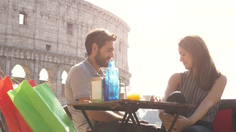 Happy-young-couple-tourists-chatting-relaxing-having-fun-sitting-at-bar-restaurant-in-front-of-colosseum-in-rome-at-sunset-with-coffee-juice-and-shopping-bags