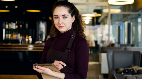 Portrait-of-cheerful-and-beautiful-young-waitress-standing,-keeping-menu,-and-smiling-sincerely-in-slylish-cafe.