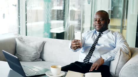 Happy-African-American-businessman-taking-a-selfie-on-his-smartphone-and-showing-a-thumb-up-gesture-while-drinking-coffee-in-modern-cafe.
