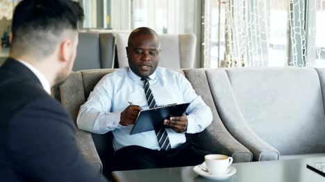 Afrikanische-amerikanische-HR-Manager-Job-interview-mit-junger-Mann-in-Anzug-und-dabei-seinen-Lebenslauf-Anwendung-im-modernen-Café-während-der-Kaffeepause