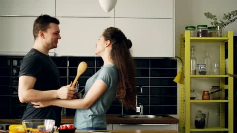 Glückliche-Paare,-die-Spaß-in-der-Küche-mit-großen-Löffeln-Fechten-und-umarmen-einander-beim-Kochen-Frühstück-zu-Hause