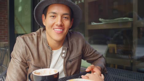 Young-man-sitting-outside-a-coffee-shop-smiles-and-holds-his-girlfriend's-hand,-portrait