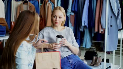 Young-woman-sitting-in-clothes-shop,-holding-coffee-and-talking-to-her-female-friend.-Ladies-having-rest-after-going-shopping.