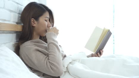 Hermosa-mujer-asiática-disfruta-de-beber-café-caliente-y-lectura-en-cama-en-su-dormitorio.-Mujer-de-Asia-usando-suéter-cómodo-sosteniendo-un-libro-y-una-taza-de-coffee.lifestyle-mujer-de-asia-en-el-concepto-de-hogar