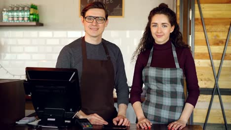 Portrait-of-two-young-good-looking-waiters-standing-at-cashier's-desk-in-coffee-house-and-smiling.-Successful-business,-happy-people-and-food-service-concept.