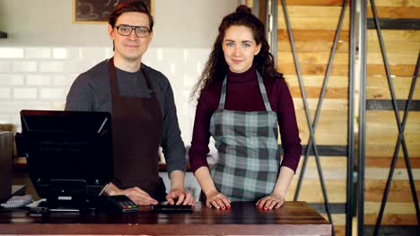 Portrait-of-two-partners-small-business-owners-standing-at-cashier's-desk-in-coffee-house-and-smiling.-Successful-business,-happy-people-and-food-service-concept.