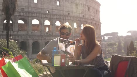 Glückliches-junges-Paar-Touristen-liest-Menü-Auswahl-von-Speisen-und-Getränken-sitzen-im-bar-Restaurant-am-Kolosseum-in-Rom-bei-Sonnenuntergang-bereit-zum-bestellen