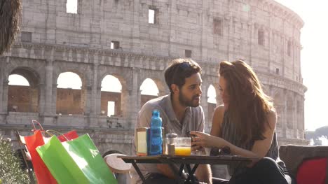 Glückliches-junges-Paar-Touristen-mit-Smartphone-sitzen-am-bar-Restaurant-am-Kolosseum-in-Rom-bei-Sonnenuntergang-mit-Kaffee-Einkaufstüten-lächelnd-mit-Spaß-SMS-durchsuchen-und-Freigeben-von-Bildern