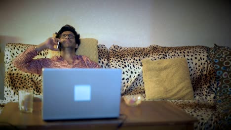 Hombre-indio-en-gafas-hablando-por-teléfono,-sentado-en-un-sofá-delante-de-un-ordenador-portátil