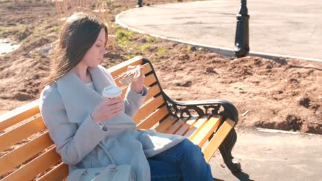 Junge-Brünette-Frau-in-Mantel-sitzt-auf-der-Bank-im-Stadtpark,-Brötchen-mit-Zucker-isst-und-trinkt-Kaffee.-Frühling.