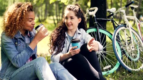 Studentinnen-sind-im-Chat-und-Kaffee-zum-Mitnehmen-im-Park-ausruhen-auf-dem-Rasen-nach-dem-Fahrrad-fahren.-Attraktive-Girls-werden-Lächeln-und-lachen-genießen-Getränke.